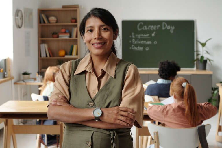 Happy young Hispanic female teacher in casualwear looking at camera with smile against intercultural schoolkids