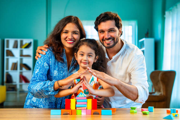 Indian Asian young married couple enjoying quality time with their adorable daughter, building a home using colorful wooden block toys at a table in their modern home